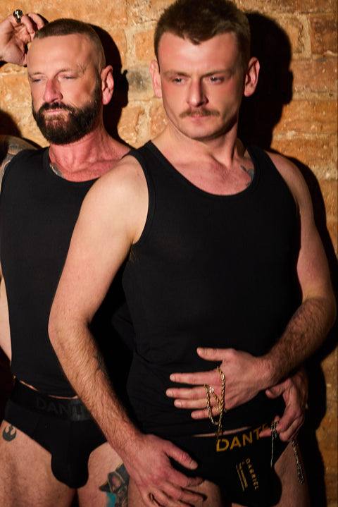 Two men wearing black tank tops against a brick wall.